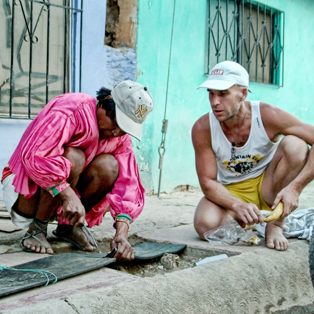 Manuel Luna crouched on a village street cutting a tyre tread into a sandal sole while Barefoot Ted watches from beside him, Copper Canyons, 2006.