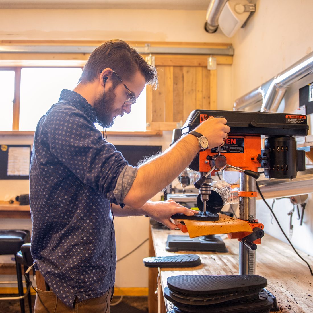 A maker at a drill press in the LUNA workshop punching a hole through a sandal footbed, cut soles stacked on the bench beside him.