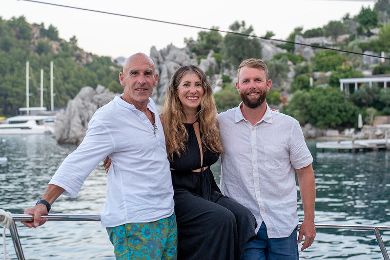 Barefoot Ted, Liv McDonald and Scott Smuin standing together at the rail of a boat, a rocky green shoreline and moored yachts behind them.