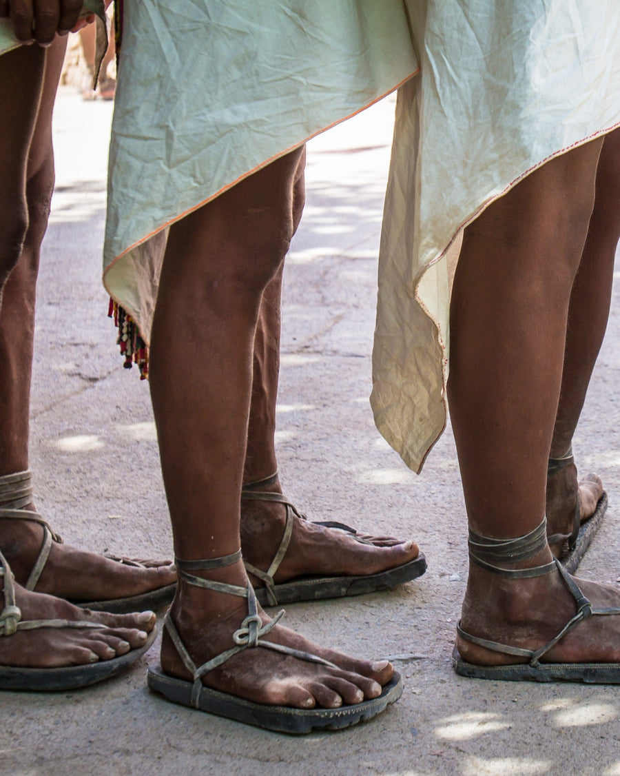 Three pairs of Tarahumara feet in traditional tire huaraches, laced at the ankle, below white cotton skirts on a dusty road.