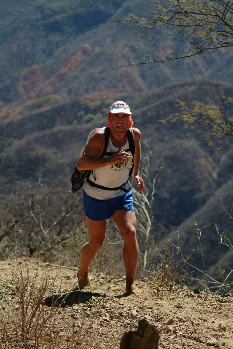 Copper Canyons, 2006. Barefoot Ted running up a steep, stony canyon trail with no shoes on, in a white cap and a Wild Mountain Run vest, the canyon wall rising behind him.