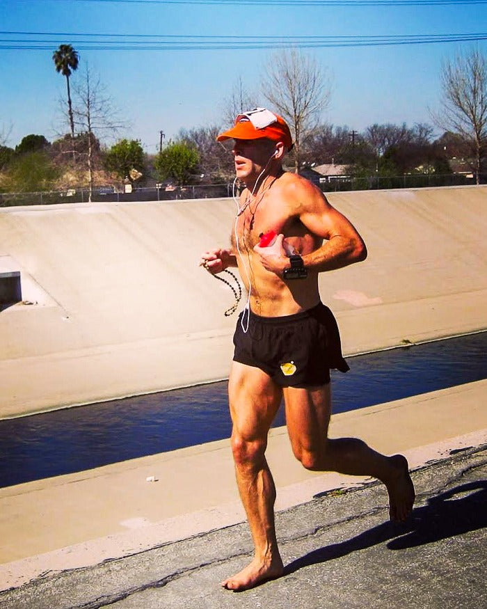 Barefoot Ted running barefoot along the sloping concrete bank of a flood channel, in an orange cap and black shorts, palm trees and houses beyond the far wall.