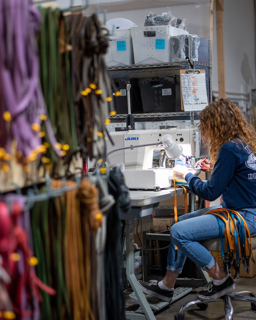 The LUNA workshop in Wenatchee, 2021. A wall of coloured laces hanging in the foreground, and a maker at a Juki sewing machine under a work light with a bundle of orange laces across her lap.