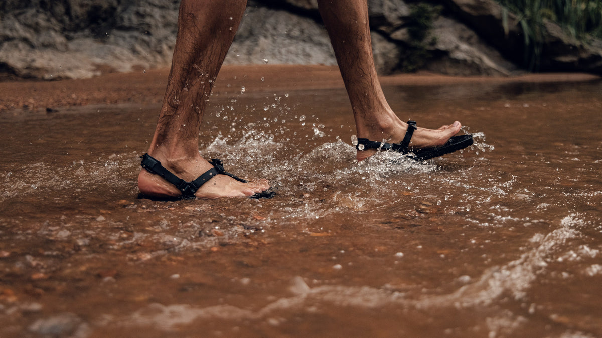Feet in black LUNA sandals mid stride, splashing through a shallow red canyon river.