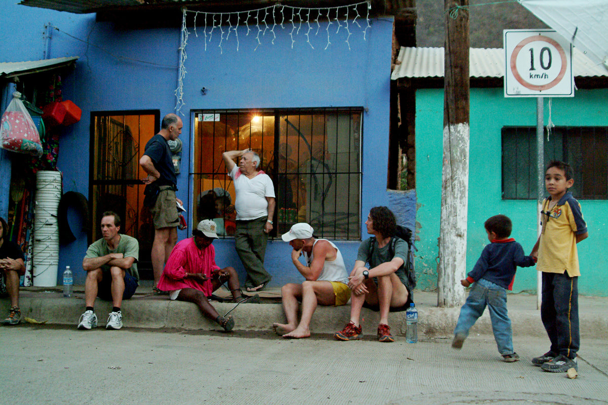 Urique, 2006. Runners and townspeople sitting together along the kerb outside a blue and green painted shop front, waiting in the late afternoon.
