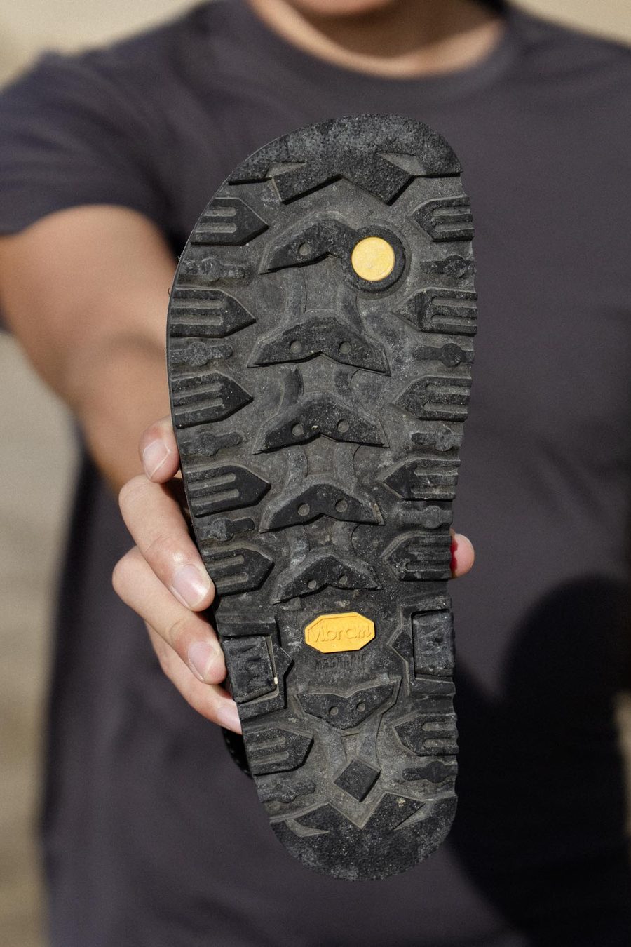 A hand holding a LUNA sandal up to the camera, the lugged Vibram outsole facing out.