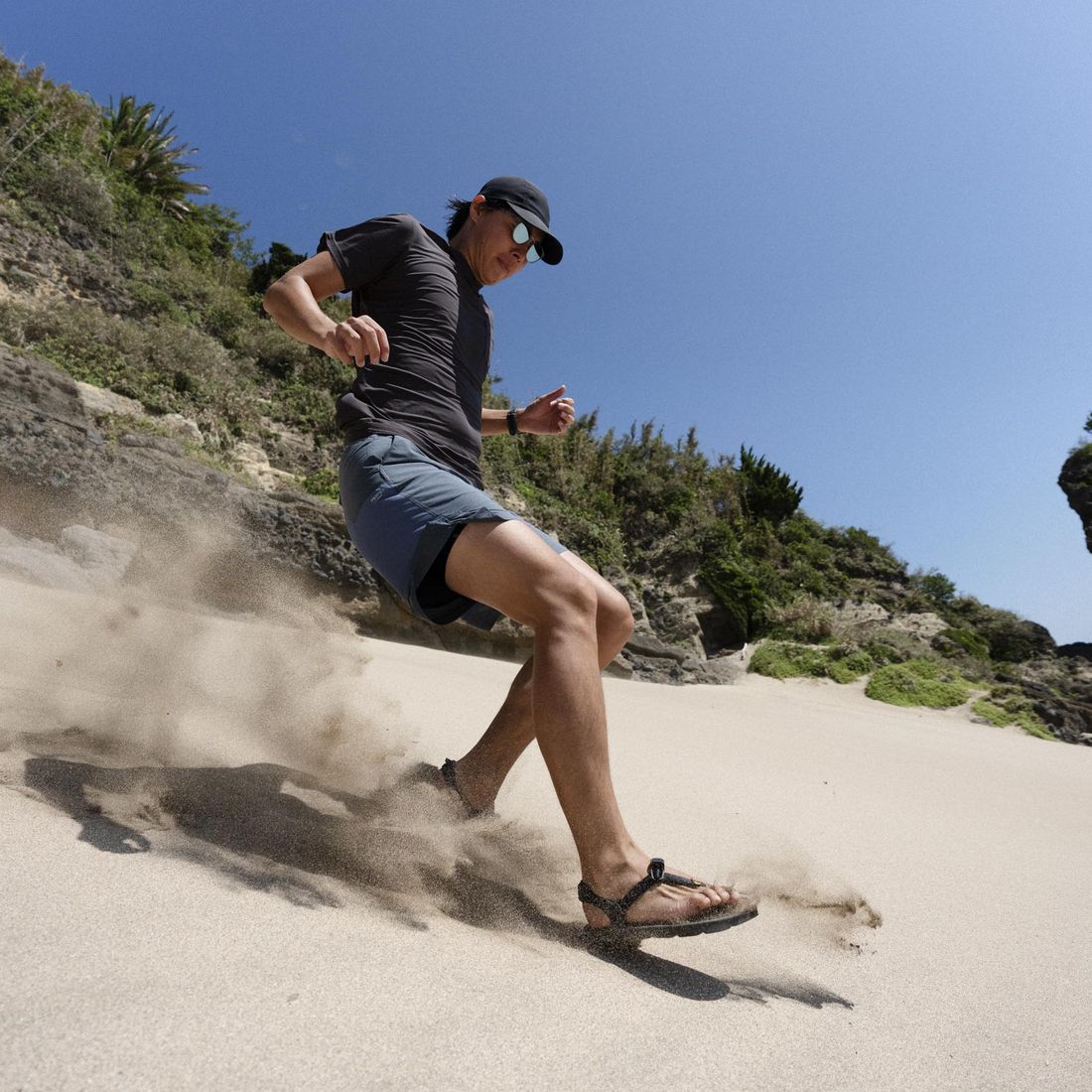 A runner in LUNAs driving up a sand slope, sand spraying behind the trailing foot, blue sky and green headland above.