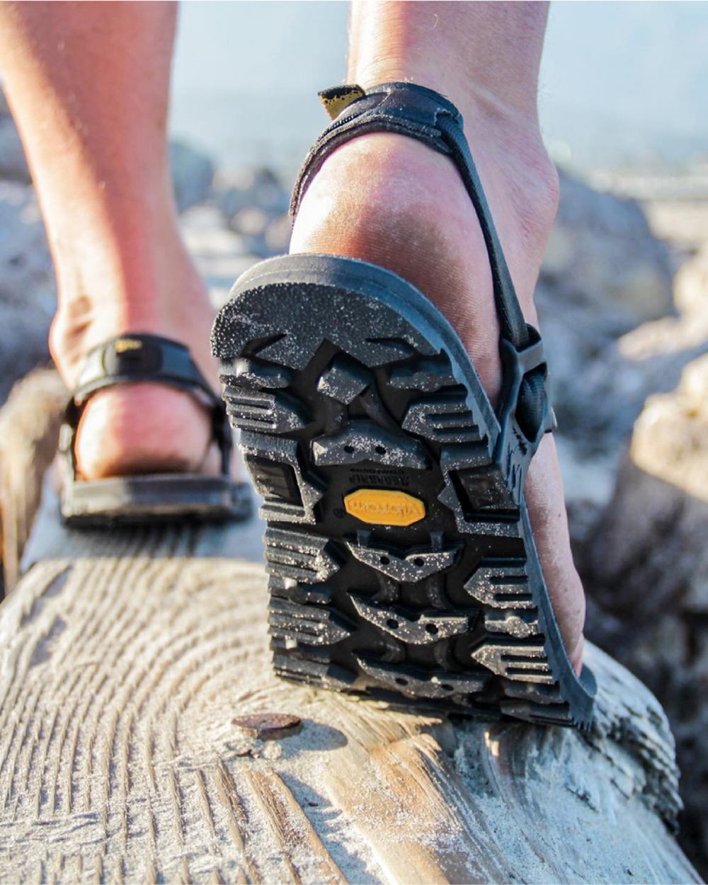 A foot in a Middle Bear sandal stepping along a fallen log, the lugged Vibram outsole turned to the camera.